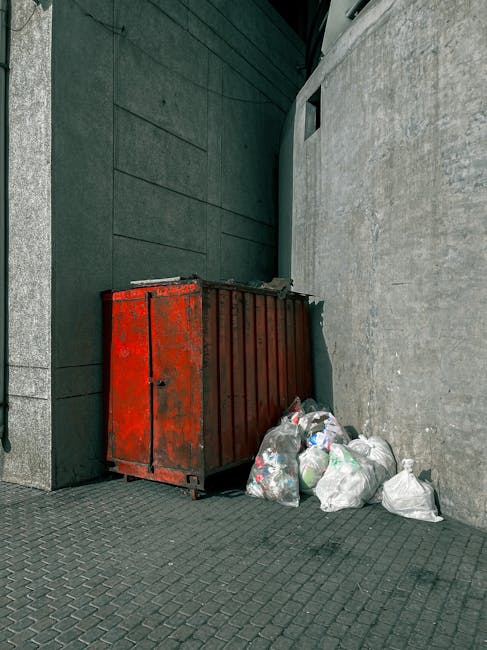 A collection of discarded cardboard boxes, some flattened and others partially intact, along with plastic bags and packaging waste, are piled up in a small outdoor corner adjacent to a brick and concr