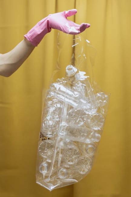 A person wearing a pink rubber glove is holding a clear plastic bag filled with crumpled and flattened transparent plastic bottles, likely used for recycling purposes. The bag is partially open at the